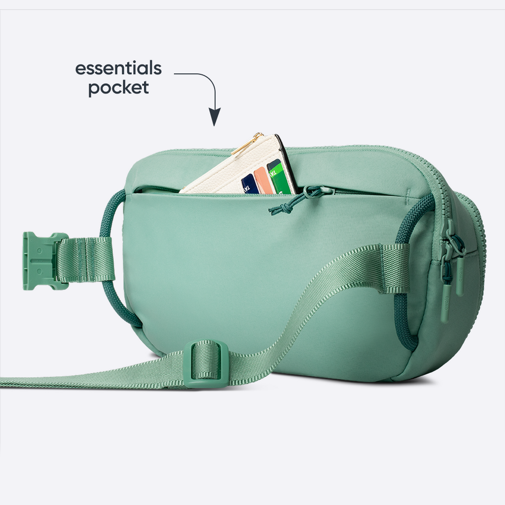 Owala All-Day Crossbody in Sea Glass, sage mint belt bag with matching strap and front zip pocket holding a card, with text reading "essentials pocket" pointing to the open pocket